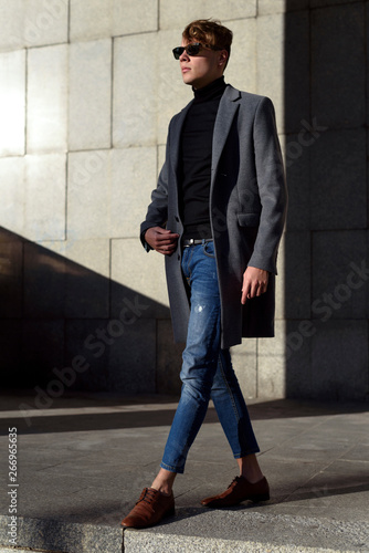 Fashionable young adult man in sunglasses. Posing in grey elegant jacket, black polo neck, denim trousers and brown mocassin shoes. Fashion trends.