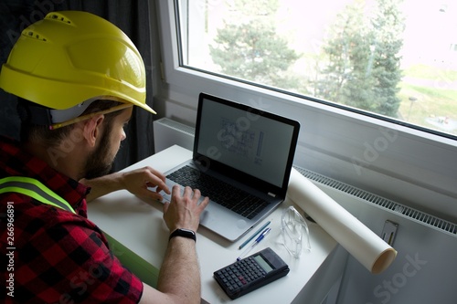 Fotografie Construction Worker is sitting at his desk at the computer, looking at building plans, counting and thinking