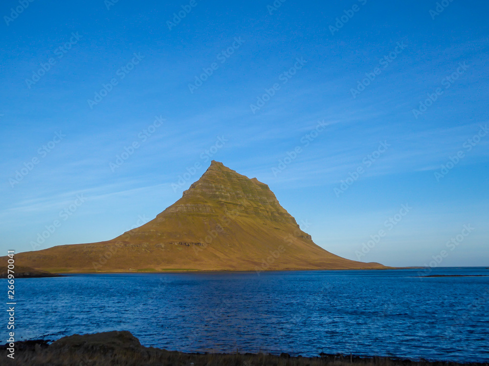 A stunning view on famous Iceland's mountain, Kirkjufell. In front of the tall hat-like mountain spreads the shallow water of a fjord. Beautiful and clear day, popular destination for a holiday.