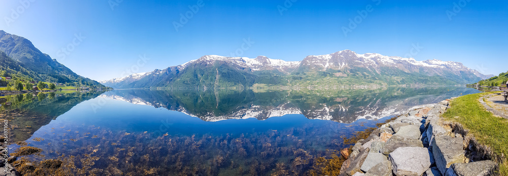 An endless chain of mountains reflecting itself in a calm water of Eidfjord. Taller parts of the mountains are partially covered with snow. Sunny and bight weather, clear blue sky. Romantic landscape - obrazy, fototapety, plakaty 