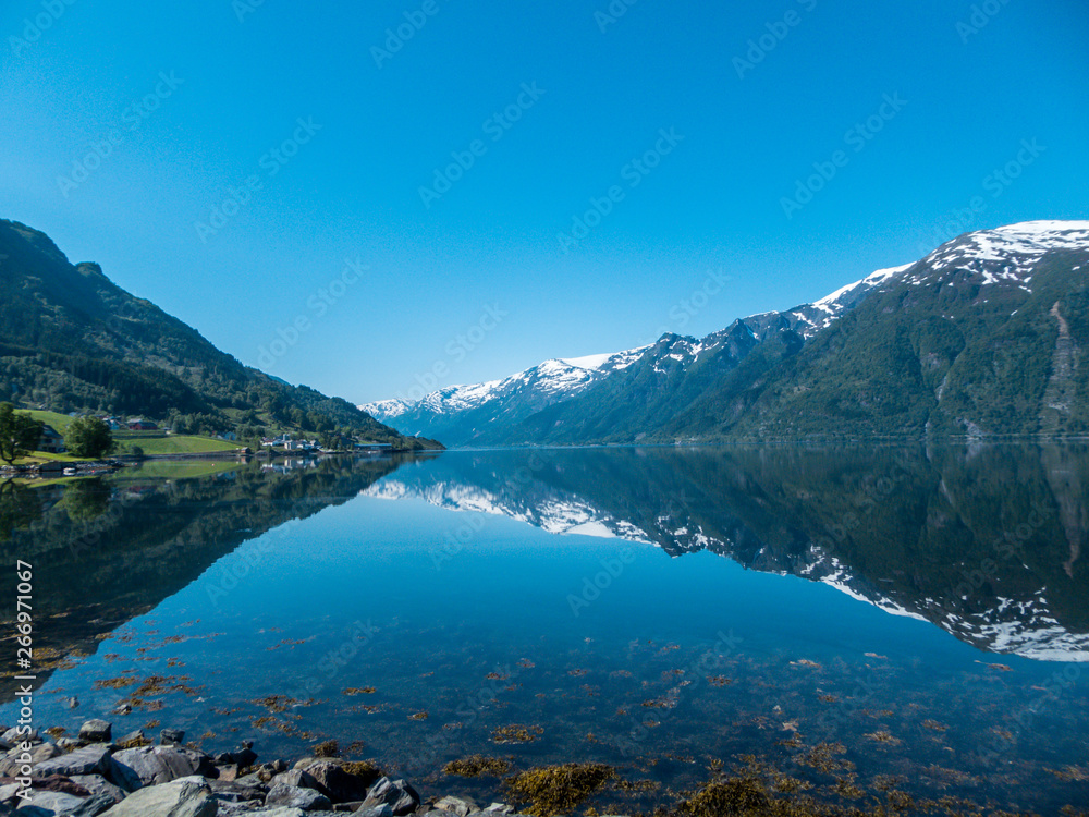 An endless chain of mountains reflecting itself in a calm water of Eidfjord. Taller parts of the mountains are partially covered with snow. Sunny and bight weather, clear blue sky. Romantic landscape