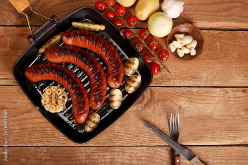 Grilled sausages with vegetables and spices in a pan on wooden background. Top view