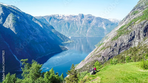 A couple standing on the meadow with a majestic view on Eidfjord from Kjeasen, Norway. Slopes of the mountains are overgrown with lush green grass. Water has dark blue color. Sunny and clear day photo