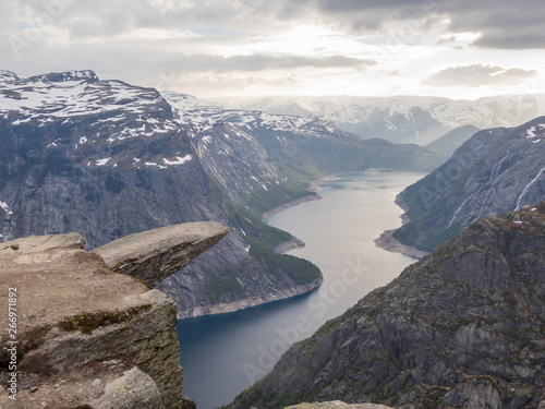 Famous rock formation, Trolltunga with a view from the above on Ringedalsvatnet lake, Norway. Rock hanging. Slopes of the mountains are partially covered with snow. The water of the lake is navy blue.