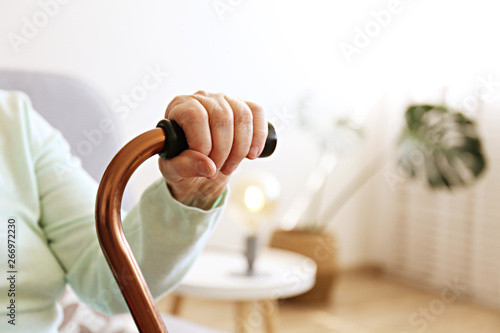 Elderly woman sitting in nursing home room holding walking quad cane with wrinked hand. Old age senior lady wearing beige cardigan, metal aid stick handle bar close up. Interior background, copy space photo