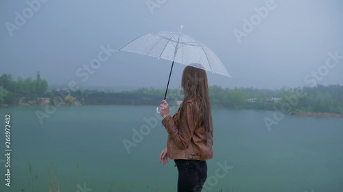 girl in a brown leather jacket with a transparent umbrella stands in the rain against the backdrop of the lake