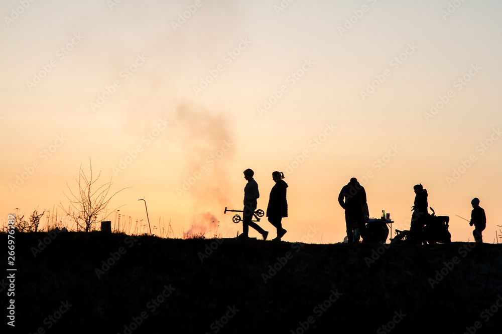 Fototapeta premium The main theme of this photo is the shadow and silhouettes of people against the backdrop of the sunset. Sunset on the bay. People relax, walk. Family roasts meat.
