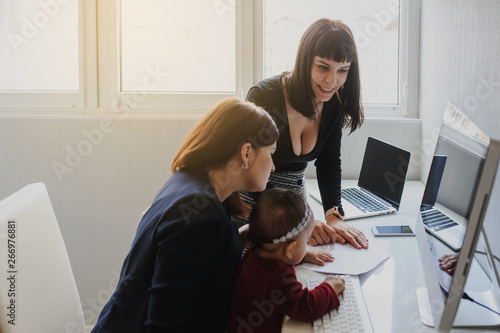 close up photo of two women in office outfit working on computers and discussing a report on the floor; one of them is with a baby girl in her arms photo