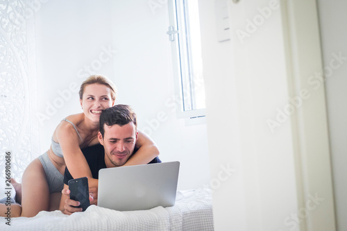 Happy young beautiful couple in playful activity at home in the bedroom onthe white bed - girl carry and hug man while he works at the laptop and phone with internet technology - lifestyle for people photo