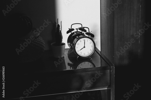 Black retro vintage alarm clock on a wooden bedside table with reflection in a polished surface in the rays of the setting sun with hard shadows