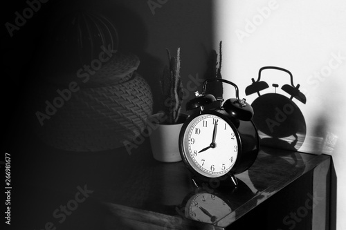 Black retro vintage alarm clock on a wooden bedside table with reflection in a polished surface in the rays of the setting sun with hard shadows