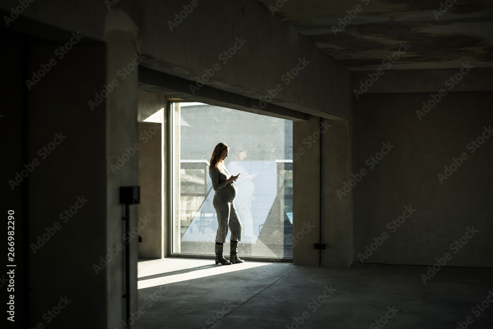Pregnant businesswoman standing at office window, using smartphone