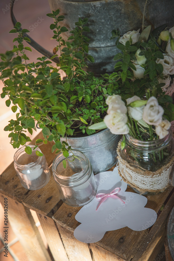 Garden composition with potted thyme plant, white roses, candles in a glass jar and a pink cardboard bear, on a wooden box in a rustic setting