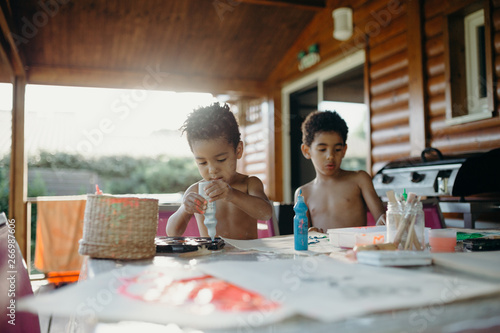 Two shirtless African American boys using bright paint to make abstract pictures on table at home photo