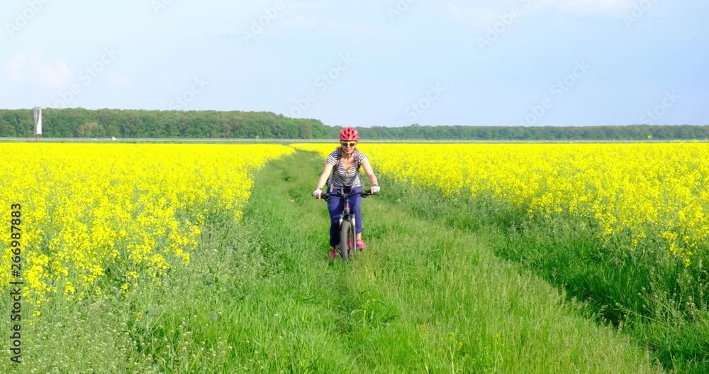 Happy woman on a mountain bike riding towards the camera, between rapeseed fields, looking up at the sun, on a trail with tall grass. Tourism in rural areas on bright springtime days. Happy woman on a mountain bike riding towards the camera, between rapeseed fields, looking up at the sun, on a trail with tall grass. Tourism in rural areas on bright springtime days.