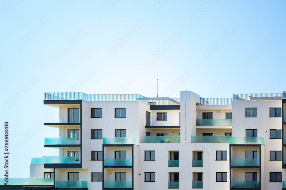 Newly built apartments building on a bright blue sky, with space for text on top. Upper part of a white building with blueish glass balconies and empty, uninhabited flats/condos, up for sale/rent.