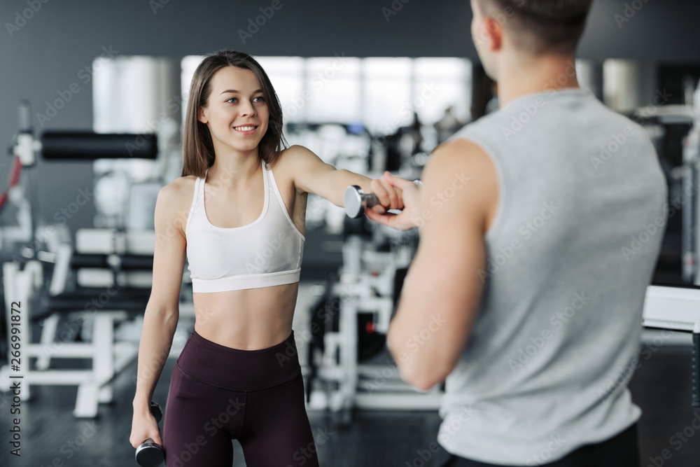 Young couple is working out at gym. Attractive woman and handsome muscular man trainer are training in light modern gym. Beautiful girl squats with dumbells under the supervision of the coach. Young couple is working out at gym. Attractive woman and handsome muscular man trainer are training in light modern gym. Beautiful girl squats with dumbells under the supervision of the coach.