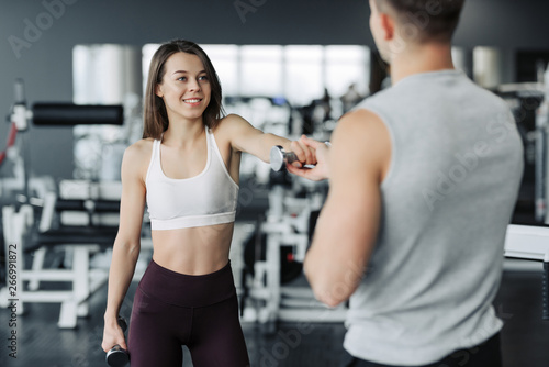 Young couple is working out at gym. Attractive woman and handsome muscular man trainer are training in light modern gym. Beautiful girl squats with dumbells under the supervision of the coach.