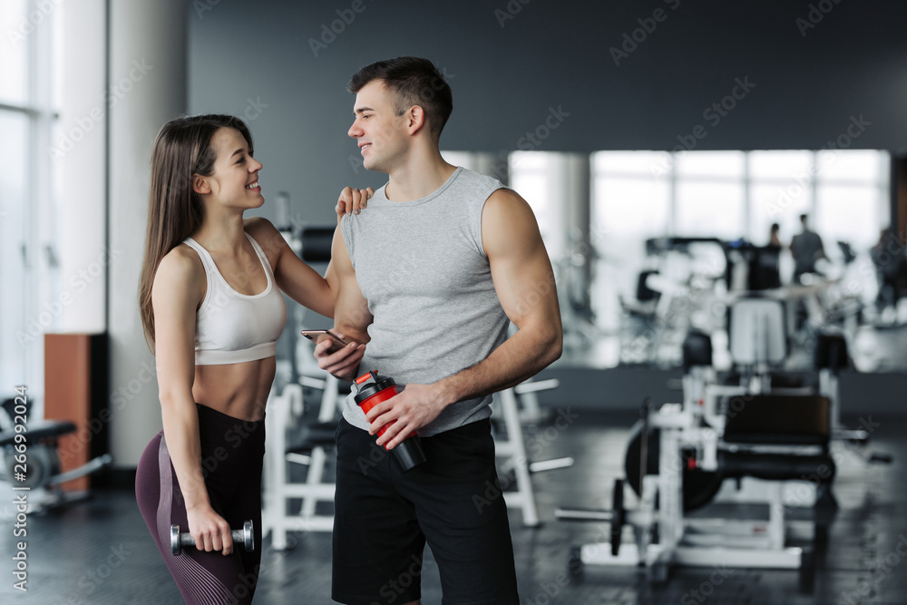 Attractive young sports people are holding bottle of water, talking and smiling while resting in gym Attractive young sports people are holding bottle of water, talking and smiling while resting in gym