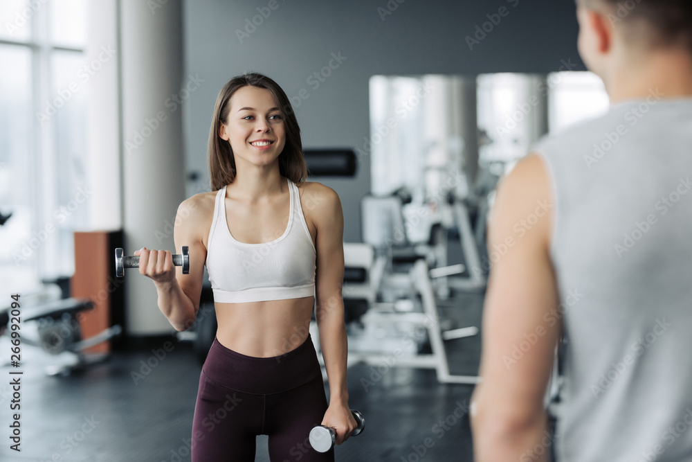 Young beautiful woman doing exercises with dumbbell in gym. Glad smiling girl is enjoying with her training process. She is working hard. Her trainer looking at this process. Young beautiful woman doing exercises with dumbbell in gym. Glad smiling girl is enjoying with her training process. She is working hard. Her trainer looking at this process.