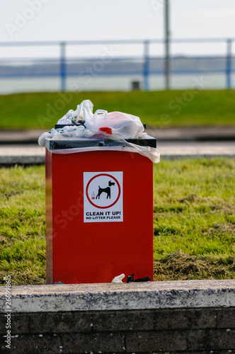 An overflowing dog waste bin by a public path