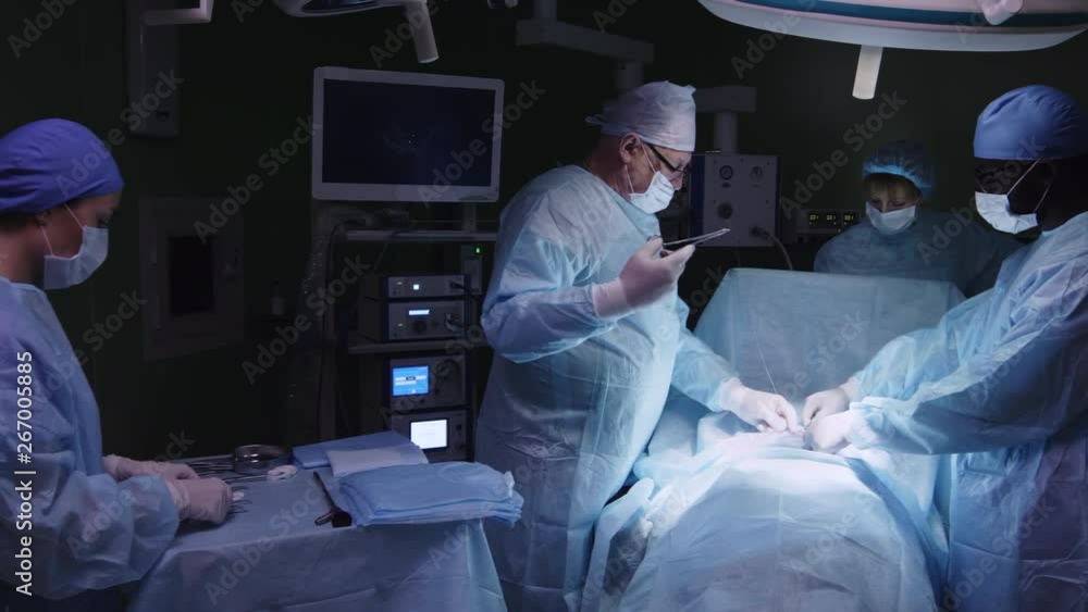 Team of experienced multiethnic surgeons operating patient and stitching incision while assistants preparing medical tools Team of experienced multiethnic surgeons operating patient and stitching incision while assistants preparing medical tools
