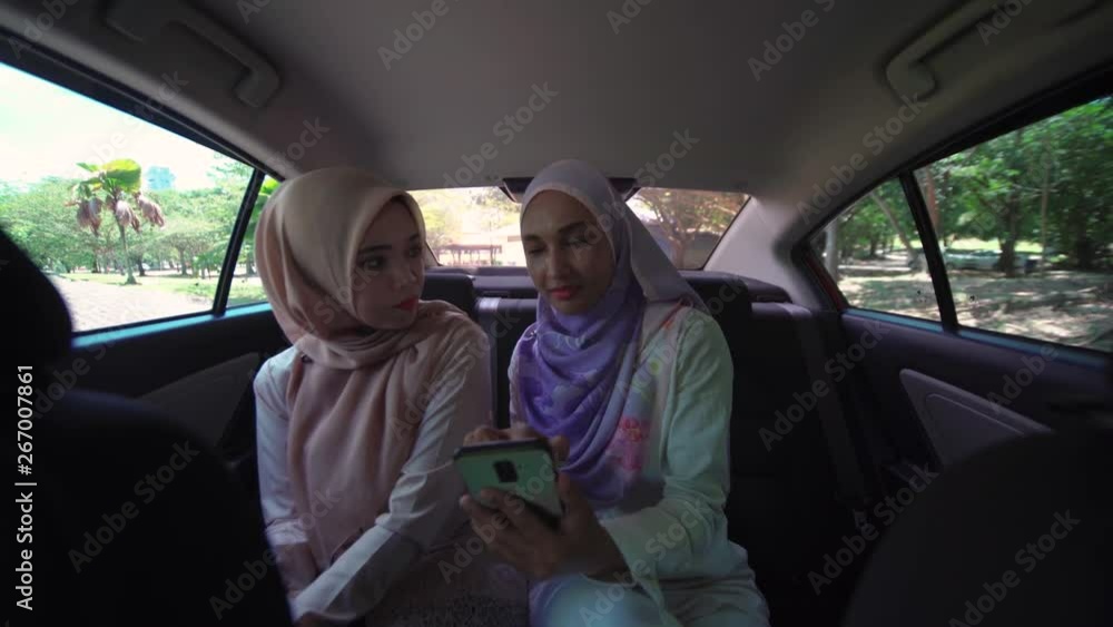 Two attractive young Asian Muslim women in hijab, talking, discussing, listening to music, looking at smartphone, at back seat of a car, sharing, friendship, transportation, ride sharing concept. Two attractive young Asian Muslim women in hijab, talking, discussing, listening to music, looking at smartphone, at back seat of a car, sharing, friendship, transportation, ride sharing concept.