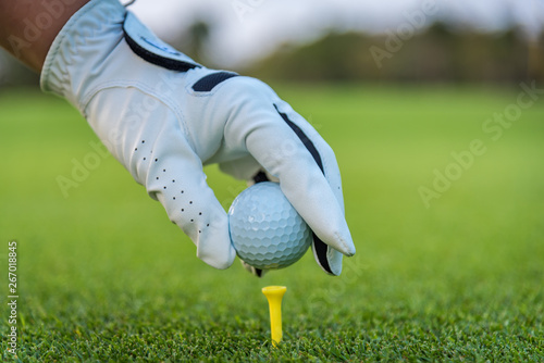 A golfer glove hand putting golf ball on tee in golf course with sunlight ray and golf course view background.
