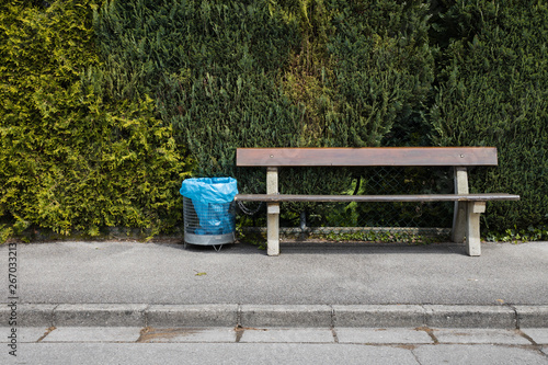 Holzbank mit angeketteter Mülltonne und blauem Müllsack vor grüner Hecke an grauer Straße