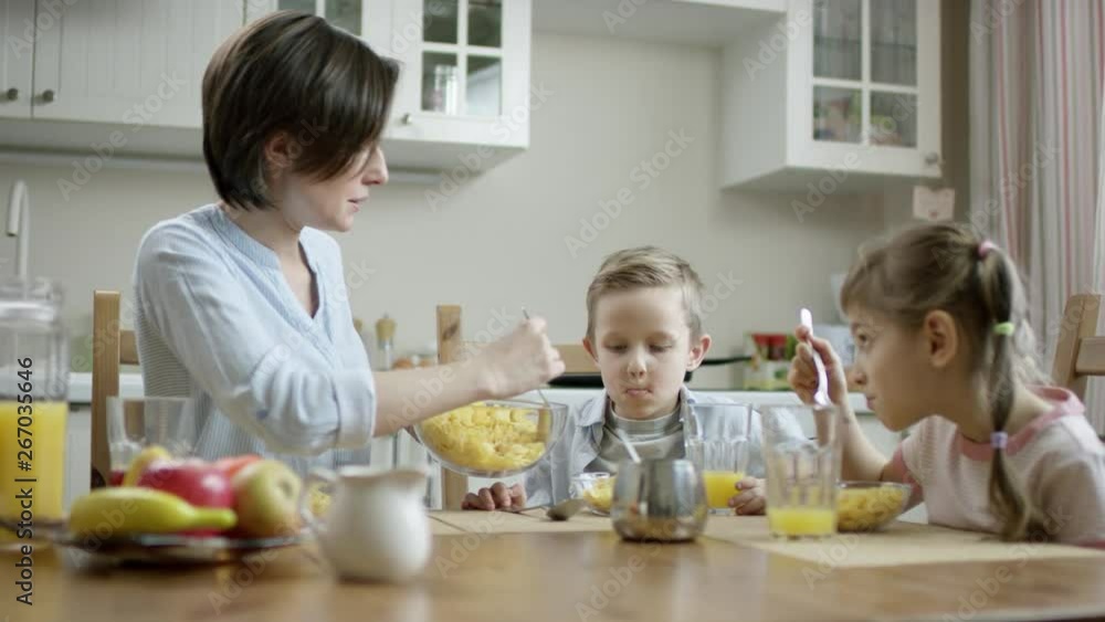 mother communicate during breakfast with her children and put cornflakes from bowl on the plate with a spoon to her son mother communicate during breakfast with her children and put cornflakes from bowl on the plate with a spoon to her son