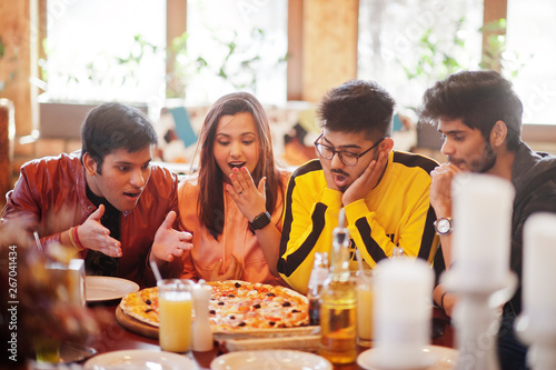 Group of asian friends eating pizza during party at pizzeria. Happy indian people having fun together, eating italian food and sitting on couch. Shocked and surprise faces.