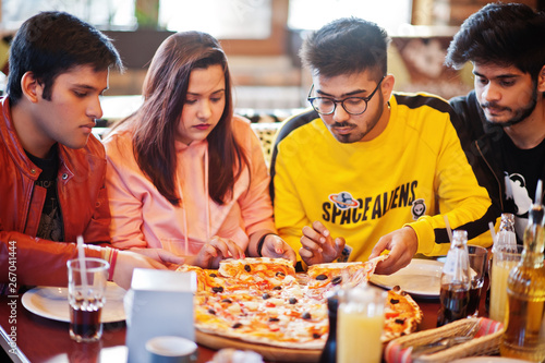 Group of asian friends eating pizza during party at pizzeria. Happy indian people having fun together  eating italian food and sitting on couch.