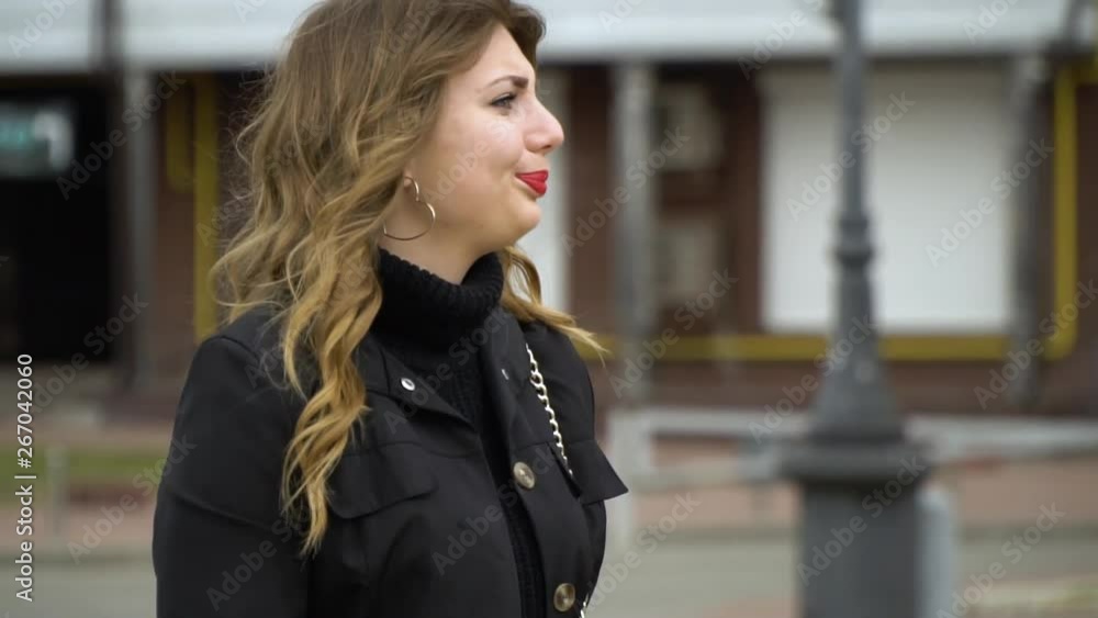 A young beautiful girl is standing on the street and waiting for someone. The girl looks around impatiently. She looks at her watch. A young beautiful girl is standing on the street and waiting for someone. The girl looks around impatiently. She looks at her watch.