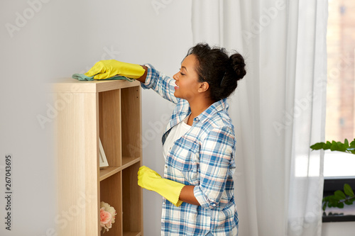people, housework and housekeeping concept - happy african american woman with dusting cloth cleaning rack at home
