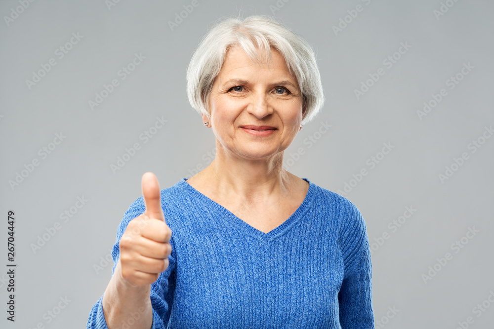 gesture and old people concept - portrait of smiling senior woman in blue sweater showing thumbs up over grey background gesture and old people concept - portrait of smiling senior woman in blue sweater showing thumbs up over grey background