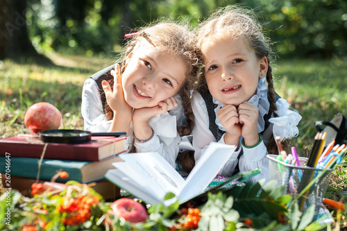 Schoolgirl girls are lying on the grass and reading books. Next to them is a globe  magnifying glass  pencils and an apple.