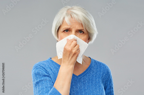 health care, flu, and hygiene concept - sick senior woman blowing nose to paper napkin over grey background