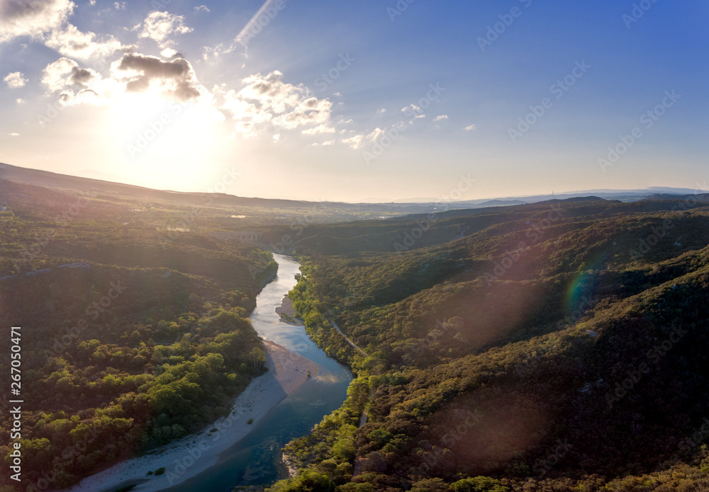 Stunning aerial drone shot of the Rhone river at sunrise. The river reflects the early morning sun and the river gorge is forested and green. An old roman bridge can be see in the distance.