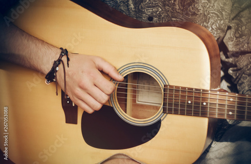 The guy in the shirt and with a bracelet on his hand plays an acoustic wooden guitar, illuminated by the sun, in the style of the 80's photo