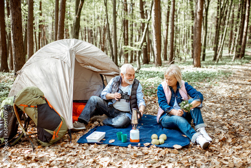Senior couple sitting together at the campsite with tent and backpacks, enjoying nature with binoculars in the forest