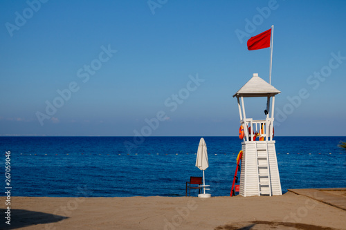 Empty lifeguard post with a red flag at a white, beach under a clear sky.