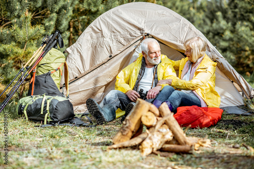 Senior couple in yellow raincoats sitting together near the tent at the campsite with fireplace in the woods