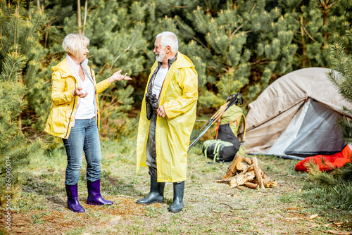 Senior couple in yellow raincoats enjoying nature at the campsite with tent and fireplace in the young pine forest