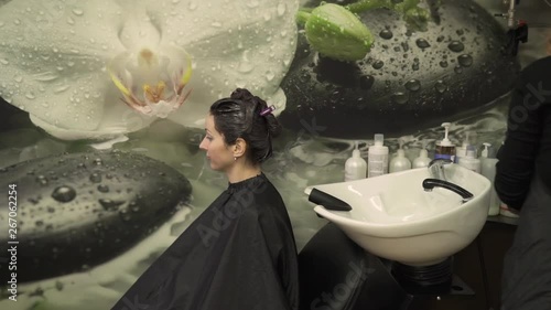 fashionable hairstylist in black apron takes off client transparent cape and starts washing long hair above special sink