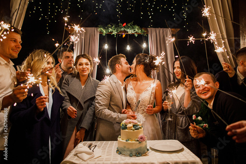 young beautiful wedding couple cut wedding cake with friends and have fun with bengal lights