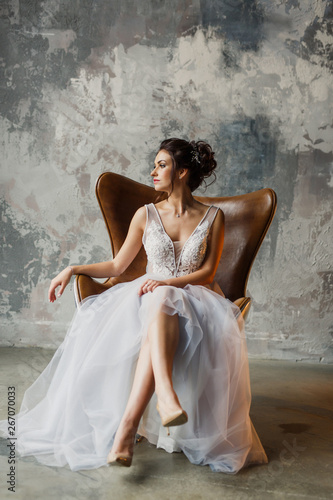 Young beautiful wedding bride in a beautiful long dress sitting in a chair in the apartment in the loft style