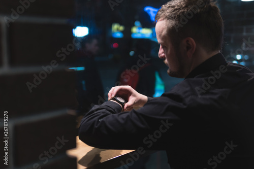 Man sits at table in cafe waiting looks at the time on the clock, night time, dark theme. Uses a smartphone and drinking coffee, learning online, marketing. Online education. window street.