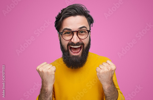 Happy bearded man wearing glasses standing with fists up on pink background. Epic win concept