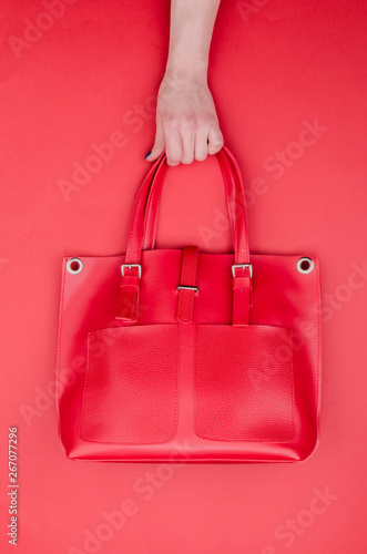 Bright summer fashion ladies accessories. Stylish red leather handbag or shopper bag on a red background. Women hands with bright manicure holding bag from top. Top View. Flat Lay. Copy Space.