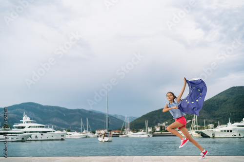 Happy child teenager girl running waving European flag at background of the sea and yachts